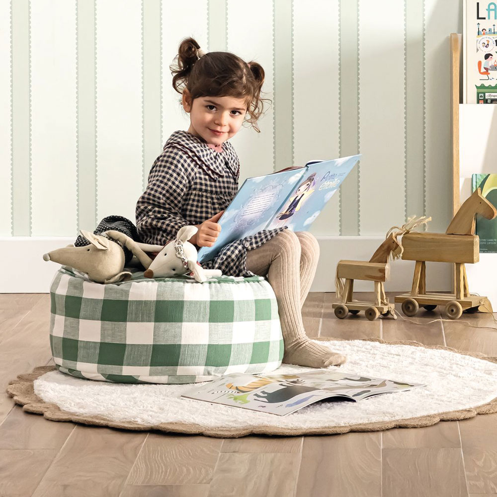 Maedchenzimmer mit zart gruener Streifentapete und Kind beim Lesen auf rundem Karo Sitzkissen