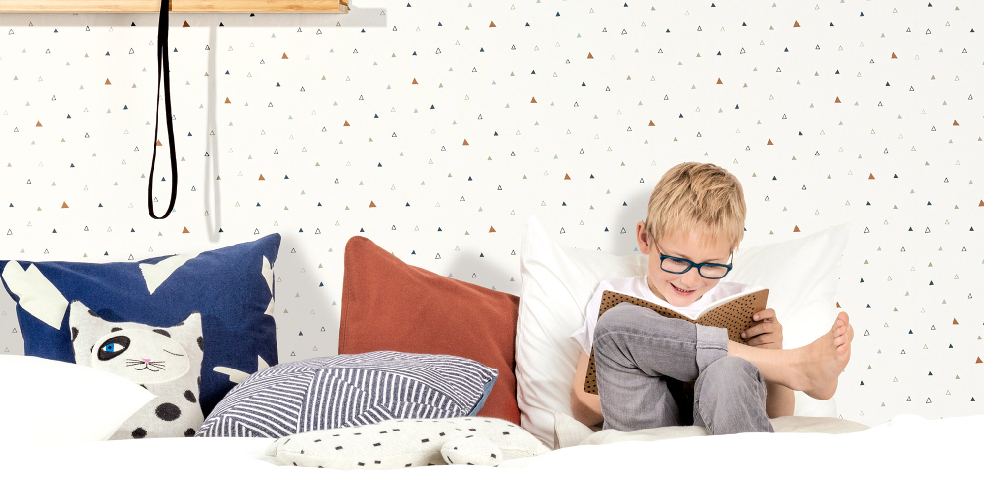 Modernes Kinderzimmer mit einer minimalistischen, weißen Tapete mit kleinen Dreiecken in Naturfarben. Ein fröhlicher Junge liest entspannt auf dem Bett.