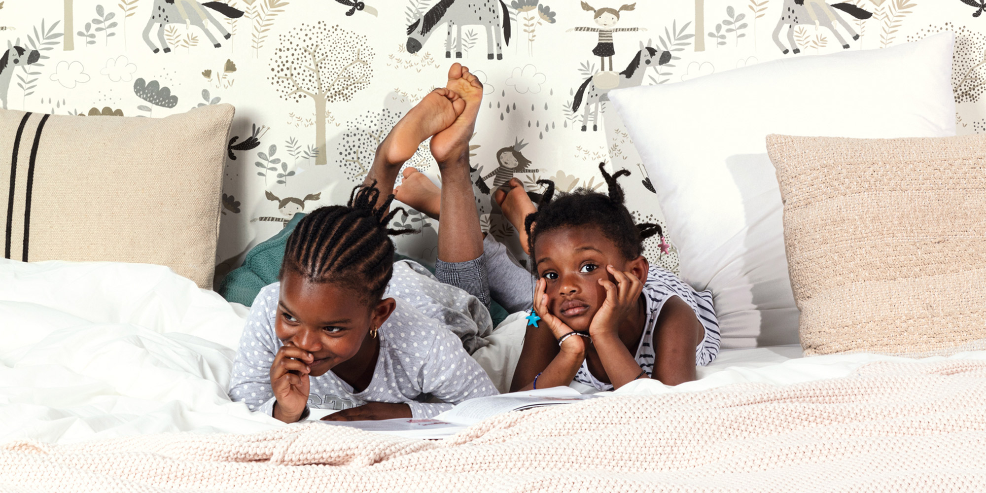 Verspielte Kinderzimmer-Tapete in Beige mit niedlichen Pferde-Illustrationen. Zwei fröhliche Kinder liegen auf dem Bett und lesen gemeinsam ein Buch.