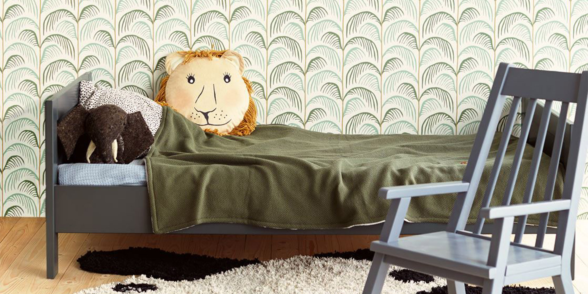 Kinderzimmer mit einer Wand hinter dem Bett, tapeziert mit einer grau-grünen Palmwedeltapete, die eine natürliche und beruhigende Atmosphäre schafft.