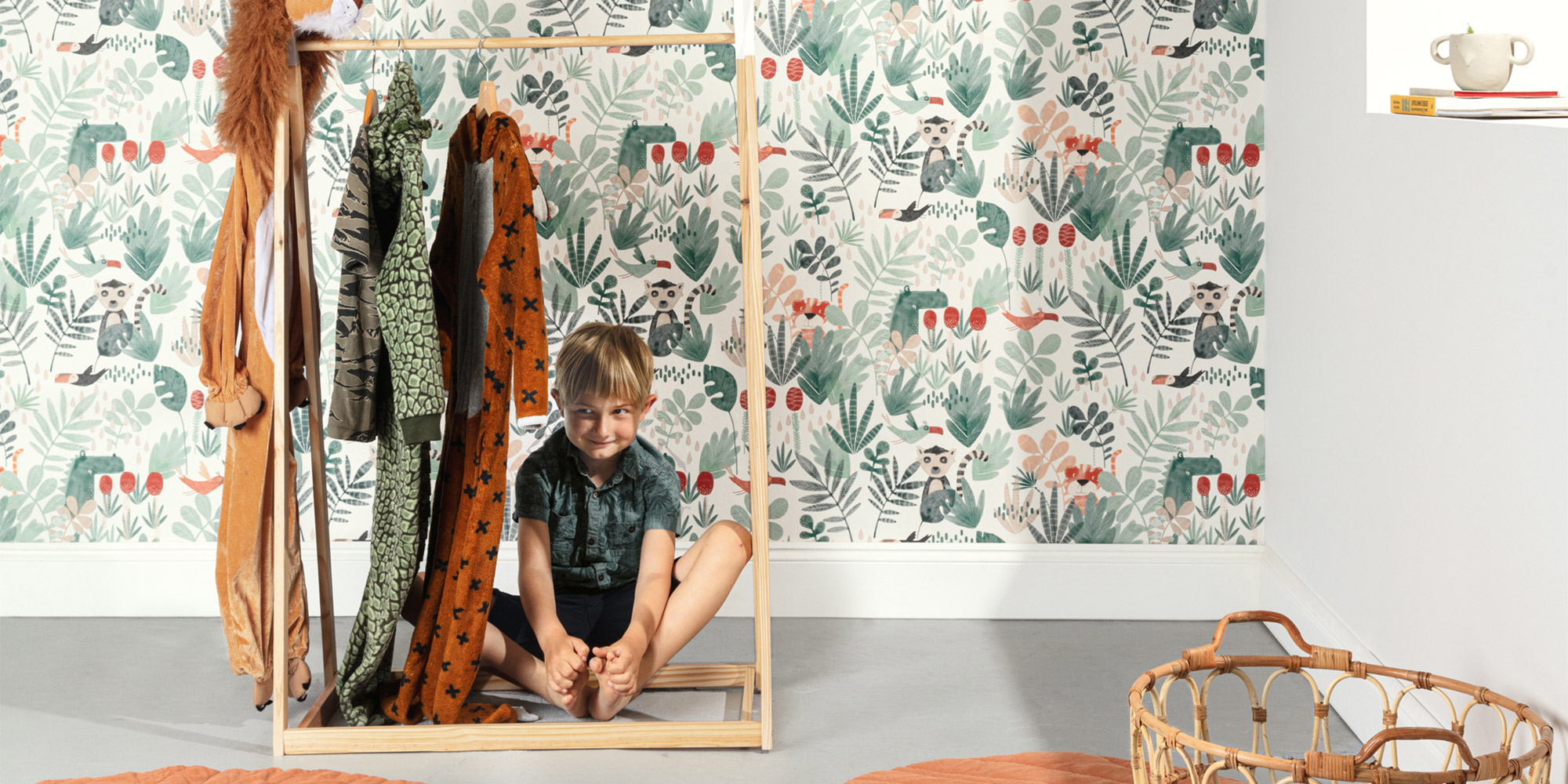 Fröhliches Kind sitzt in einem Holzgestell vor einer Kinderzimmer-Tapete mit niedlichen Lemuren, grünen Blättern und verspielten Dschungeltieren.