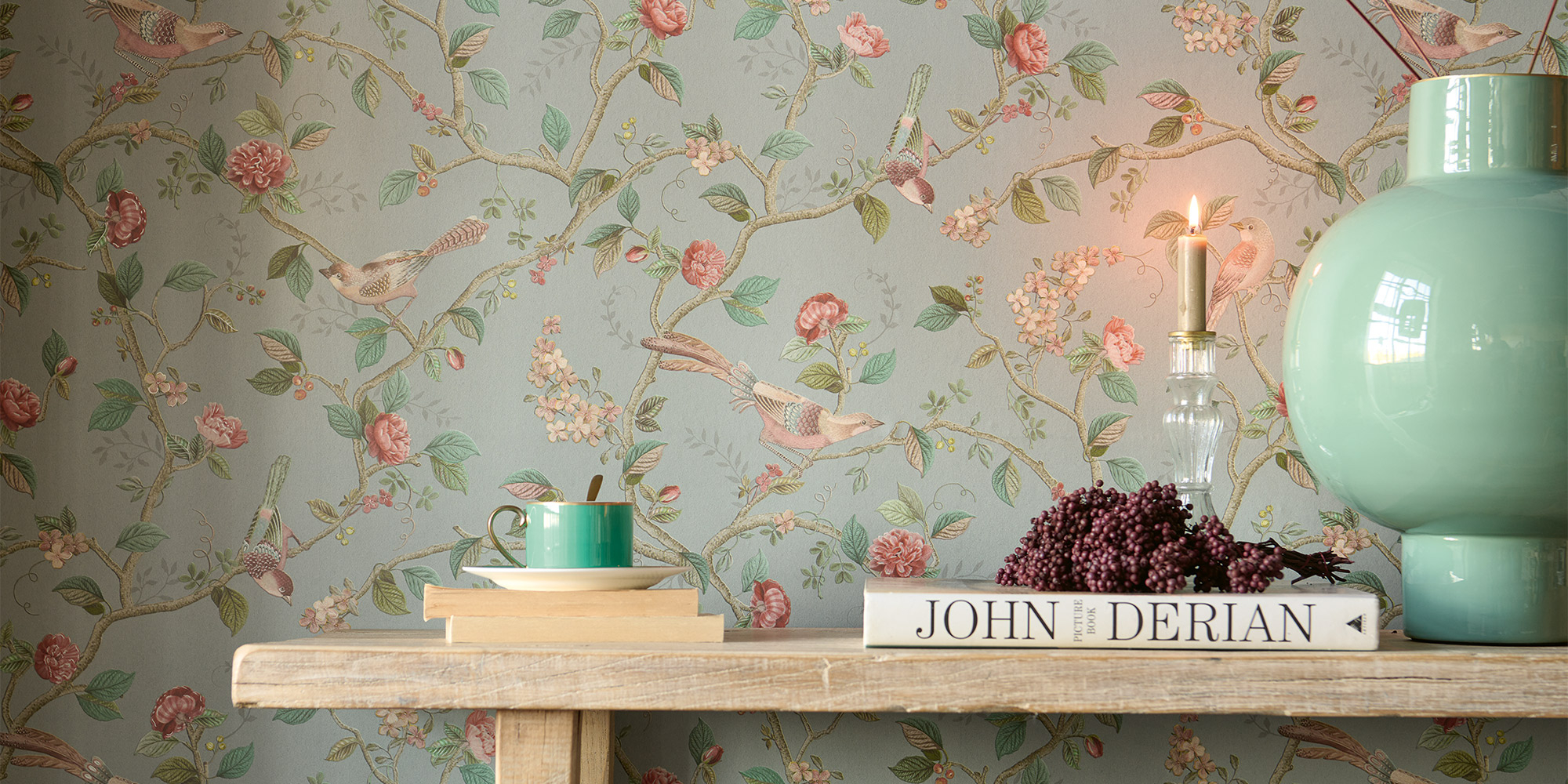 Elegante Wandtapete mit filigranen Vögeln und blühenden Ästen in Pastelltönen. Dekorative Elemente wie ein grünes Vase und ein Buch schaffen eine wohnliche Atmosphäre.