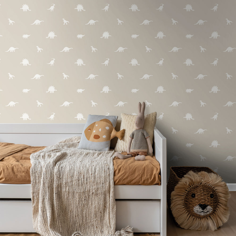Beige Dino-Tapete im Kinderzimmer mit weißen Dinosauriermotiven, Kinderbett, Kissen und Kuscheltieren als ruhige Wandgestaltung