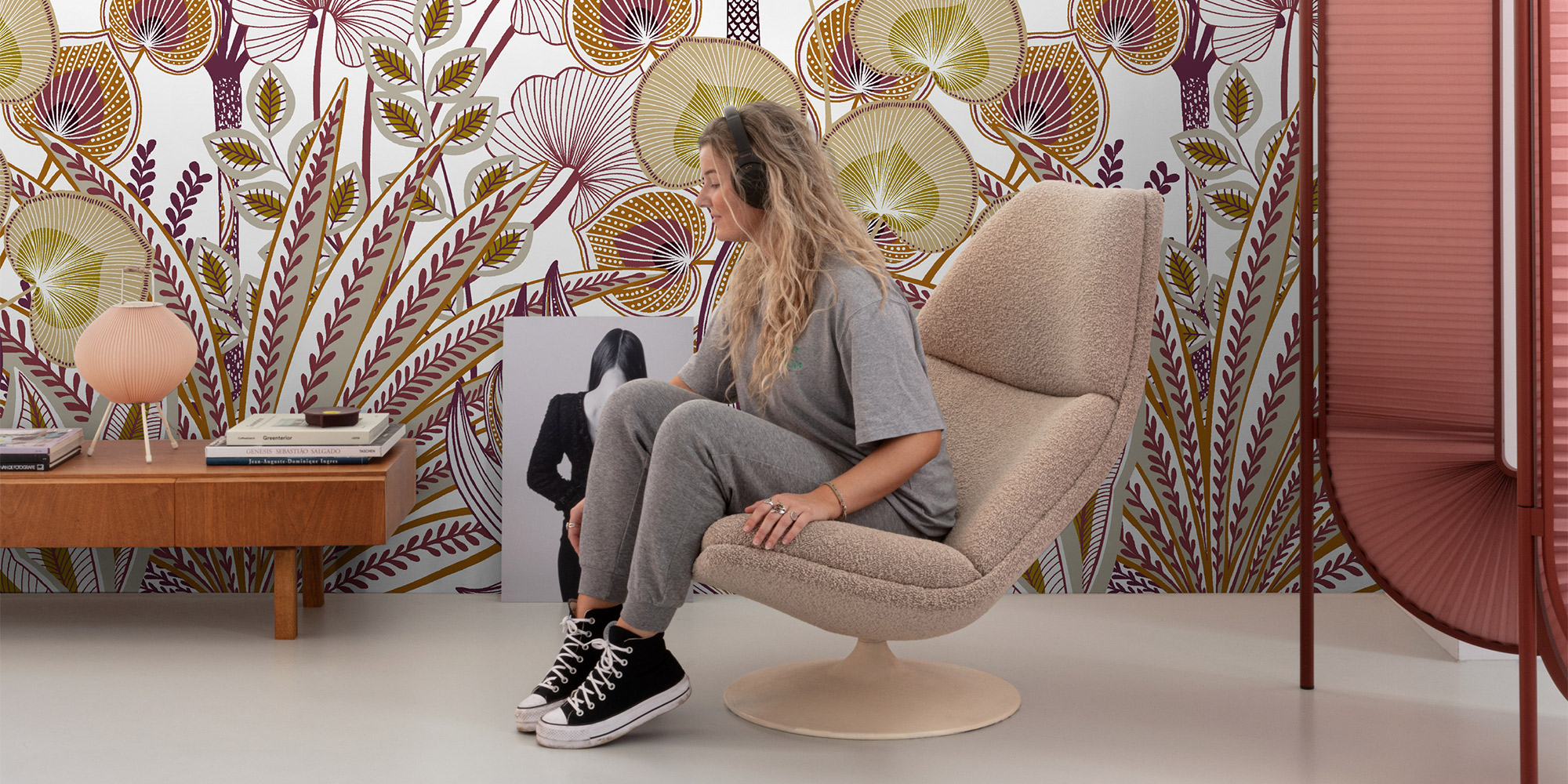 Eine junge Frau sitzt entspannt auf einem Sessel vor einem floralen Wandbild in Beige, Rot und Oliv